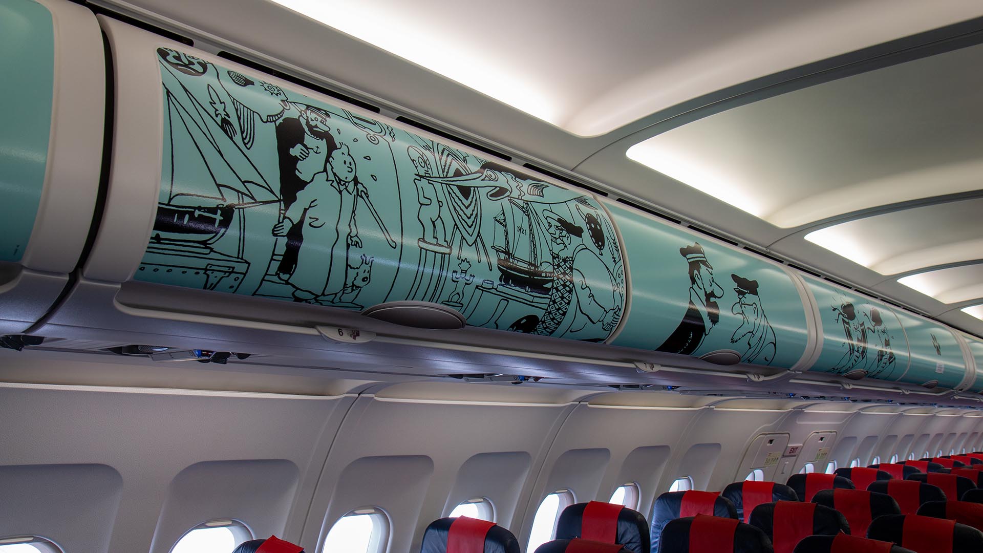 Overhead luggage bins inside an aircraft cabin decorated with a blue Tintin comic strip mural.