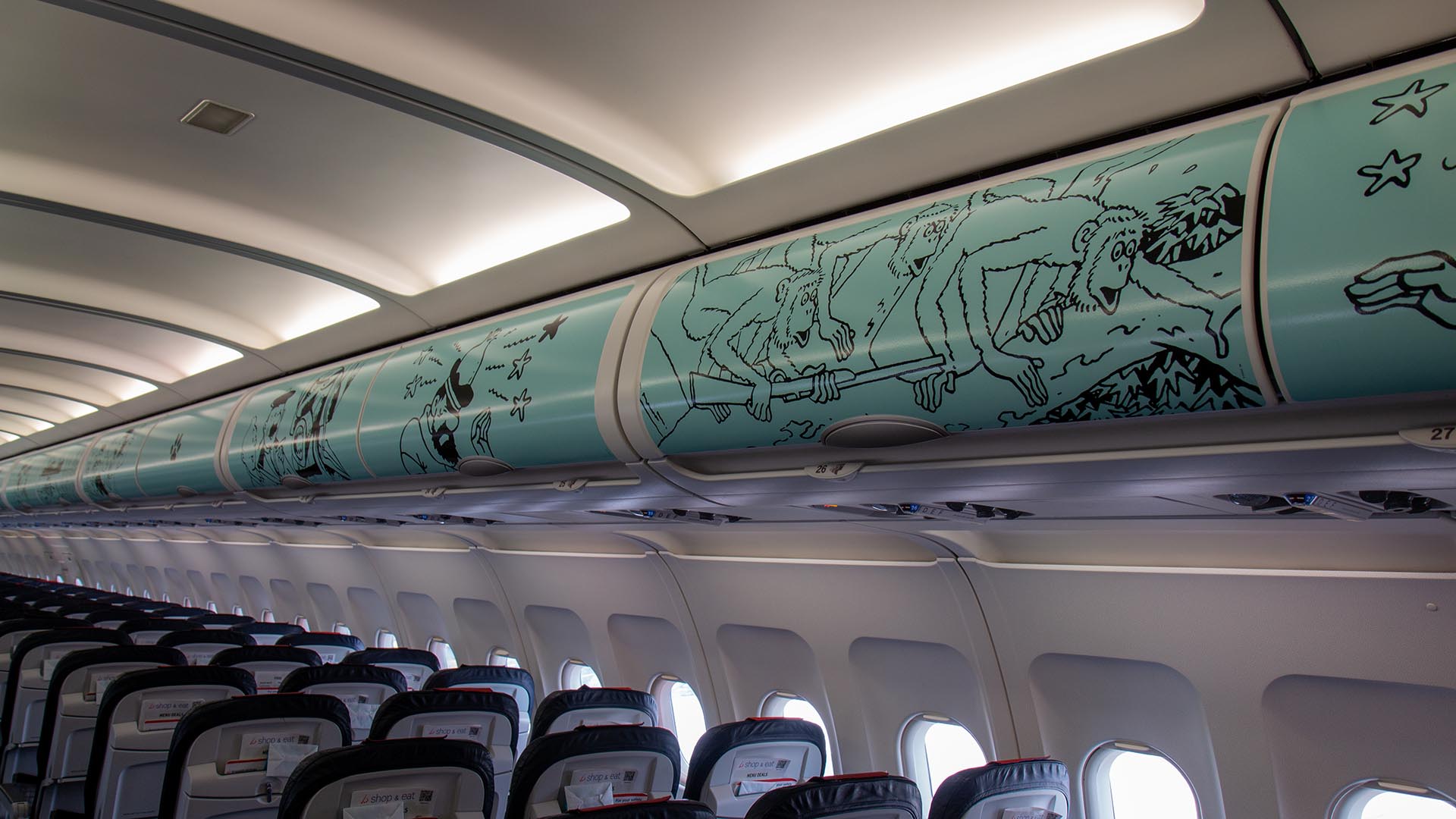 Overhead luggage bins inside an aircraft cabin decorated with a comic strip mural of monkeys.