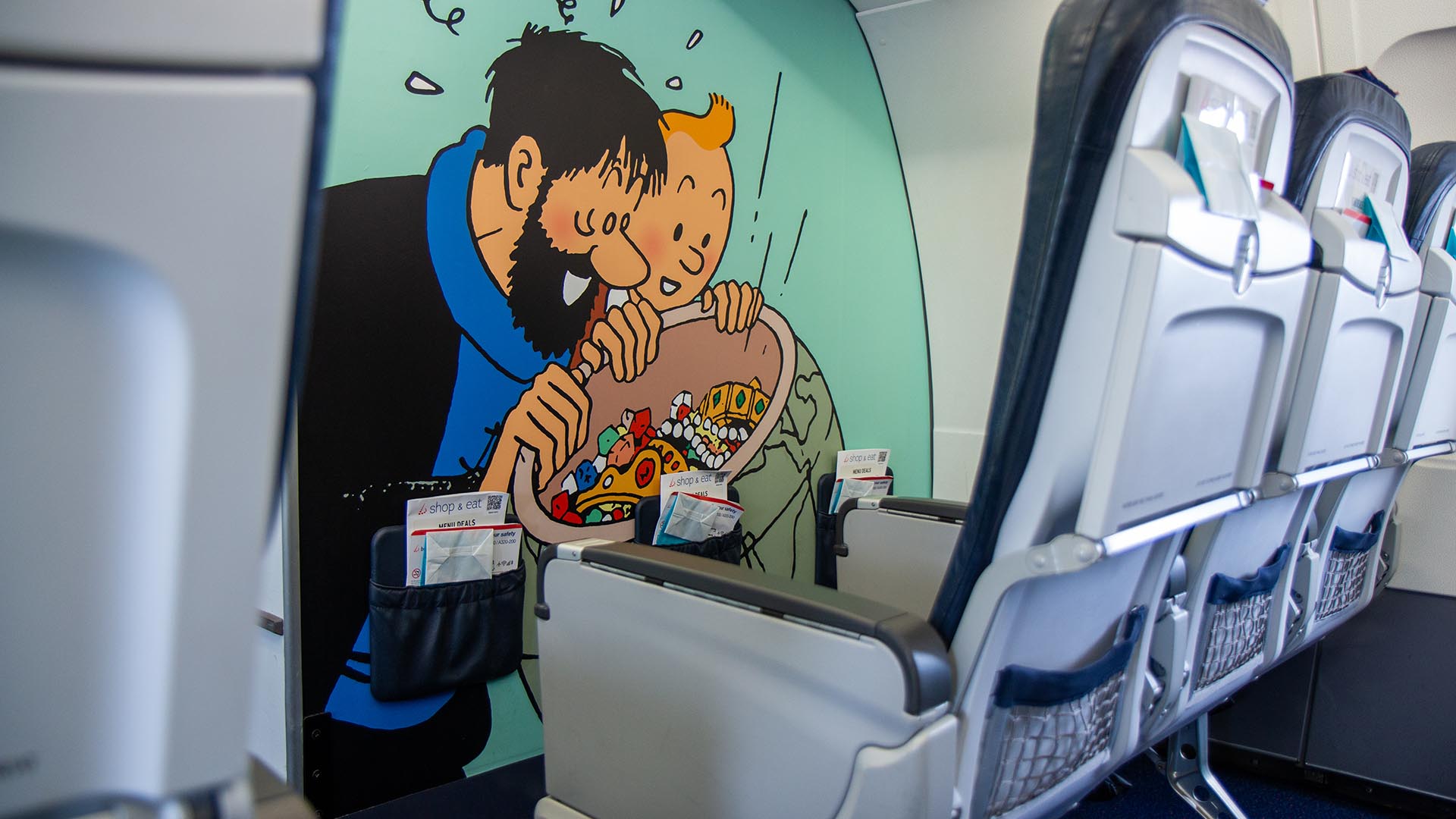 Interior of the aircraft cabin featuring a rear wall mural of Captain Haddock and Tintin looking at a pile of treasure behind the passenger seats.